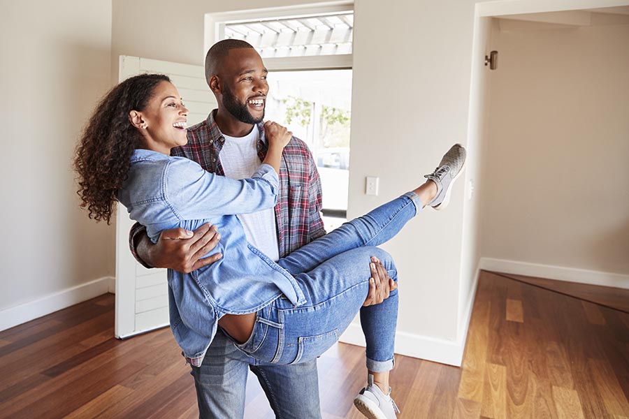 Personal Insurance - Husband Carries His Wife Into Their New, Empty Home, Both Smiling and Looking Around the House
